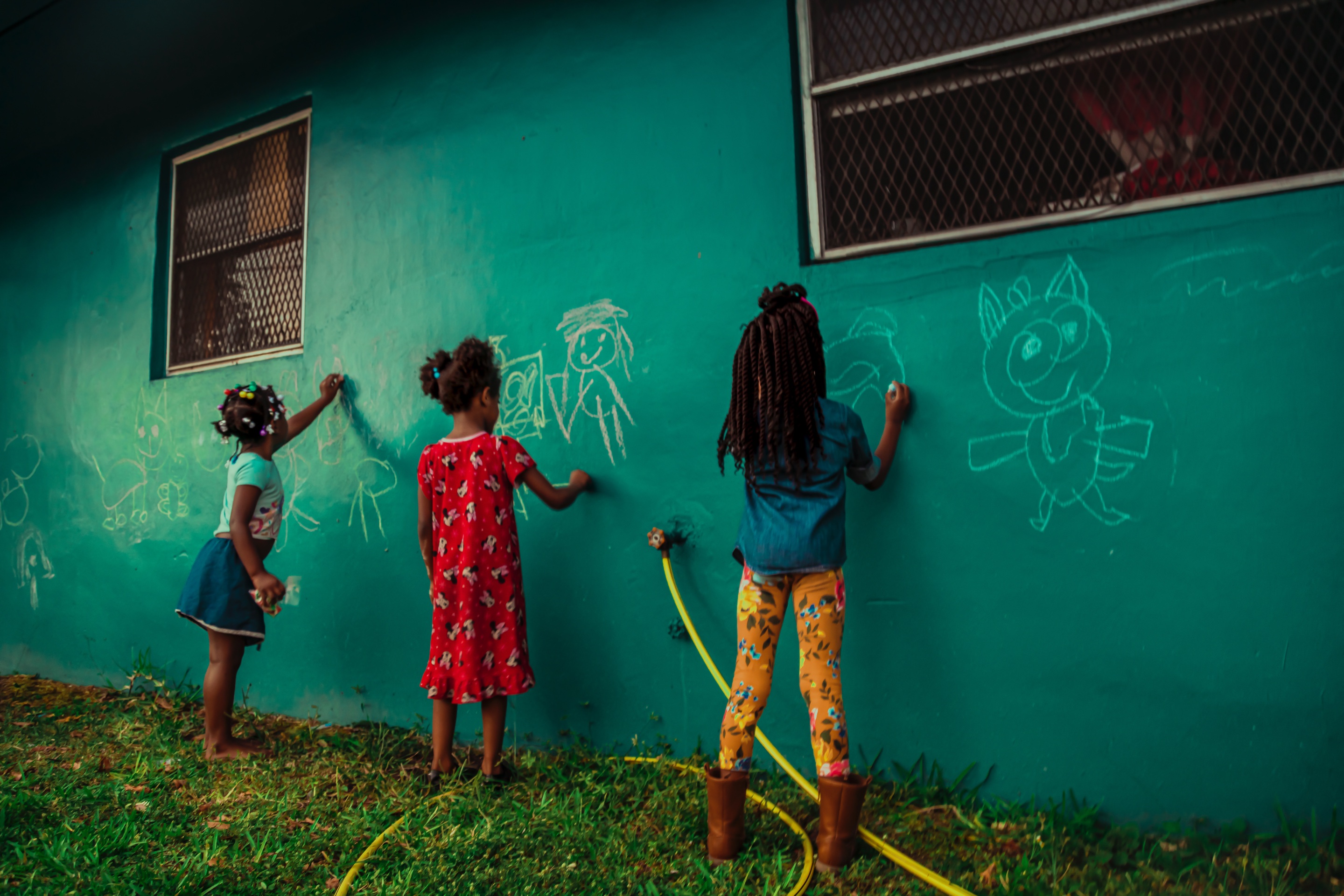 Three young girls drawing their world in chalk across a teal wall. Queens, by Jade Lilly